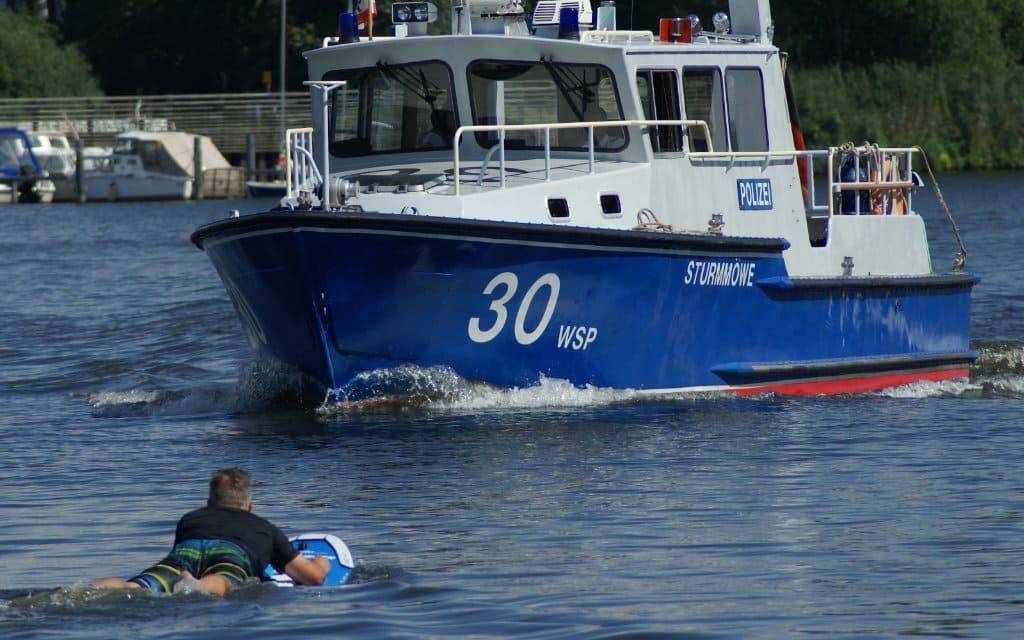 Een boot van de waterpolitie vaart langs een rijder die op zijn knieën op een eFoil-board ligt