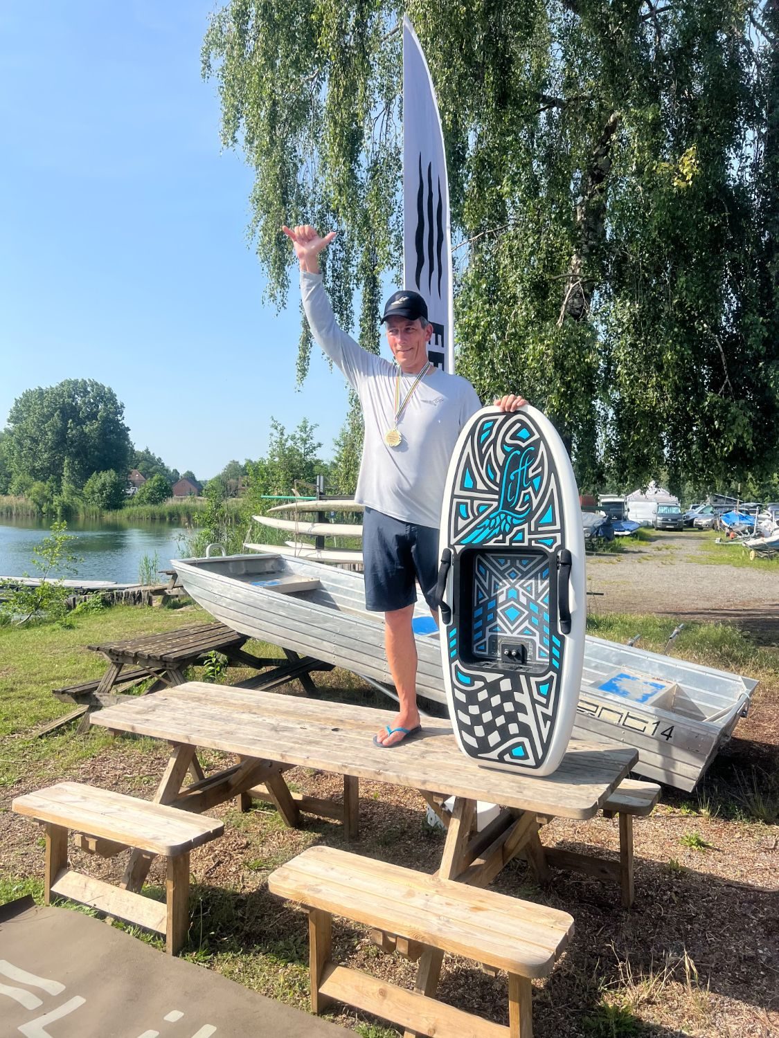 Piet Varkevisser staat met gouden medaille en zijn board op een picknicktafel aan het water en steekt zijn hand op