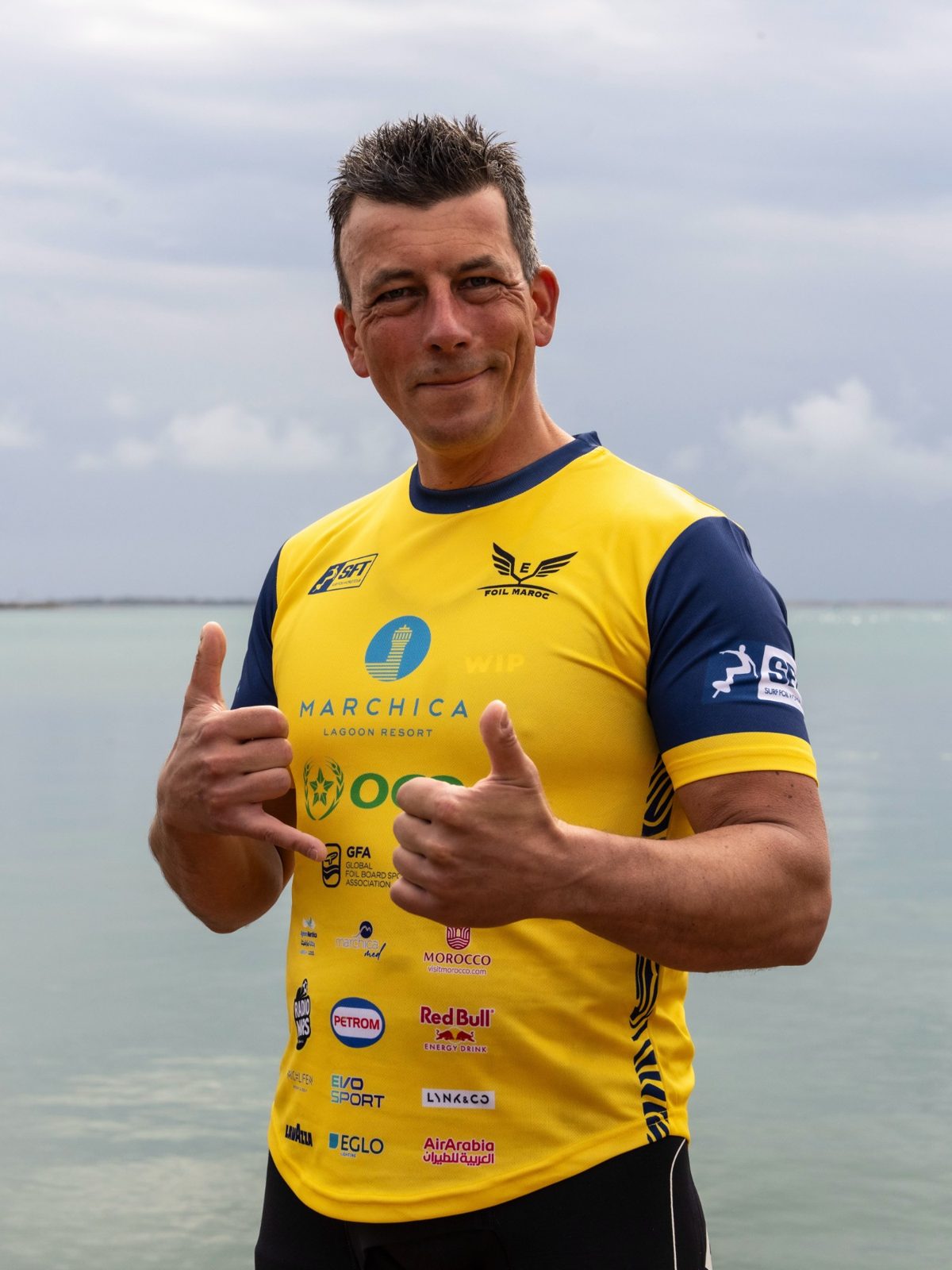 Piet Varkevisser in een geel wedstrijdshirt van de Surf Foil World Tour maakt het shaka-gebaar, met het water op de achtergrond